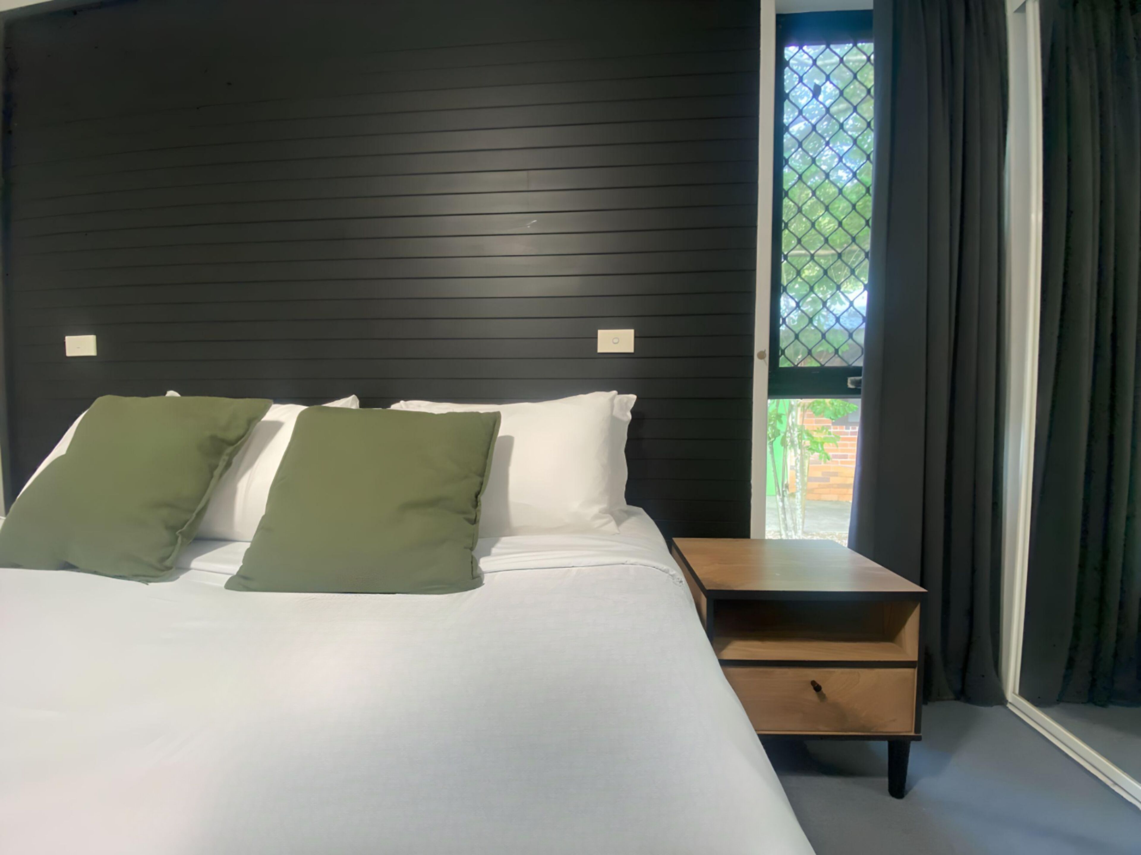 Ein Schlafzimmer mit einem Bett, zwei Kissen, einem Nachttisch aus Holz und einem Fenster mit Vorhängen.