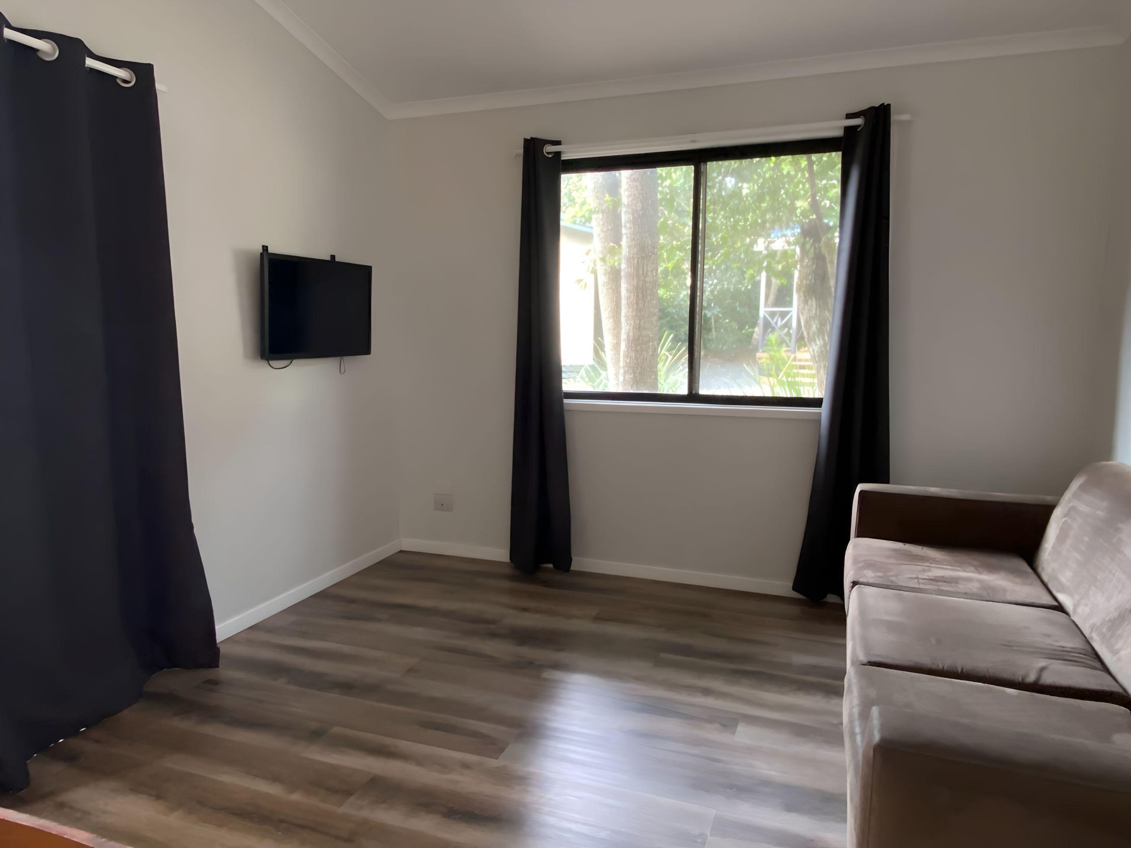 A room with a large window, a flat-screen TV mounted on the wall, and a brown sofa.