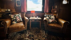 Una habitación acogedora con dos sillones de cuero, una mesita, una ventana con cortinas, una guitarra y un cuadro en la pared.