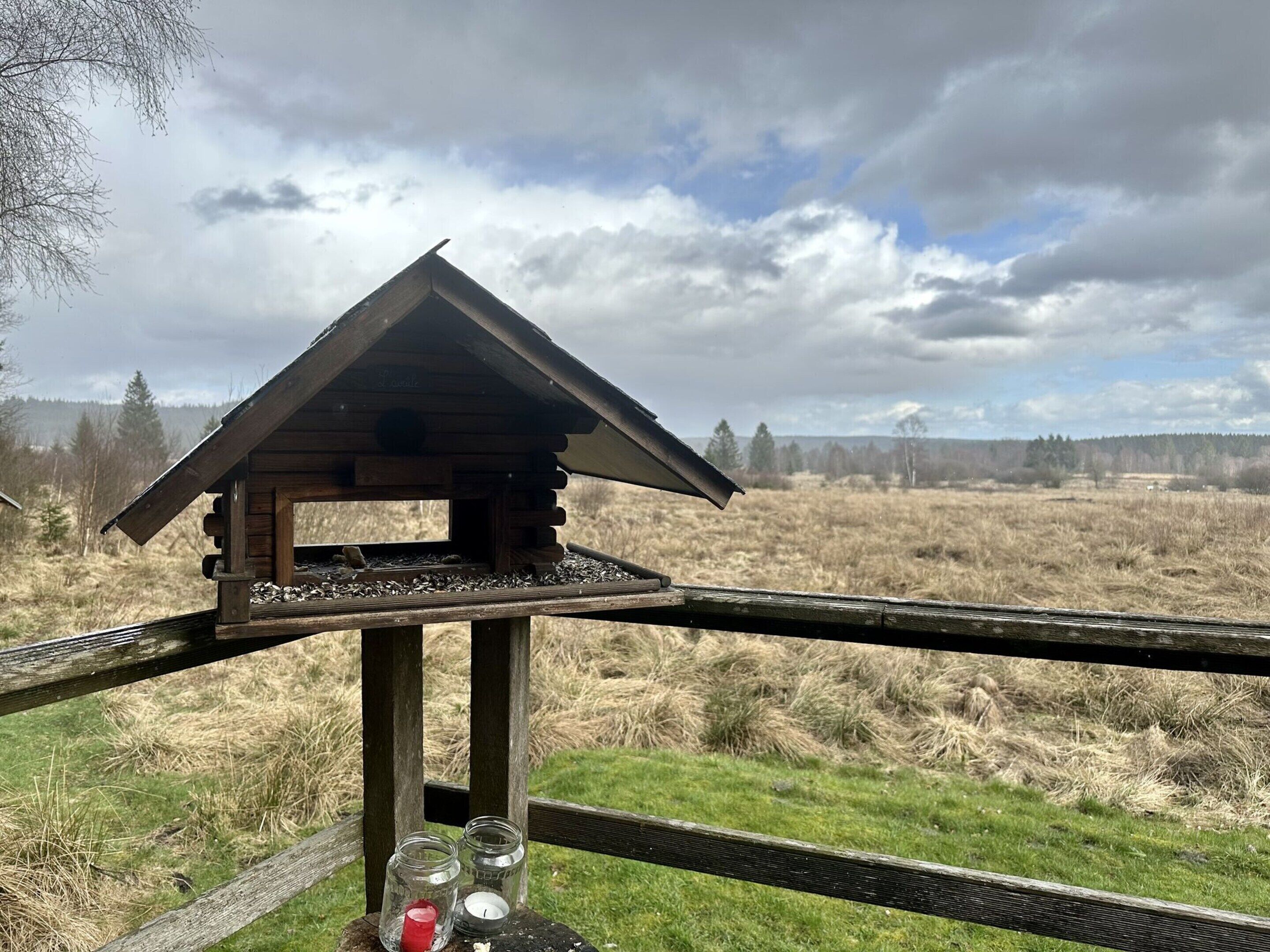 Une nichée à oiseaux en bois, posée sur une structure également en bois, avec un champ herbeux et des arbres en arrière-plan.