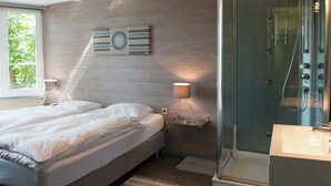 A modern bedroom with a large bed, bedside lamps, a glass shower enclosure, and a view of greenery outside. - Alluring Holiday Home in Malmedy With Sauna (Malmedy)