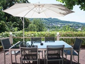 A table with four chairs and a large umbrella, set on a patio with a view of a town and hills.