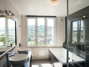 A modern bathroom with two sinks, a bathtub, and a large window offering a city view.
