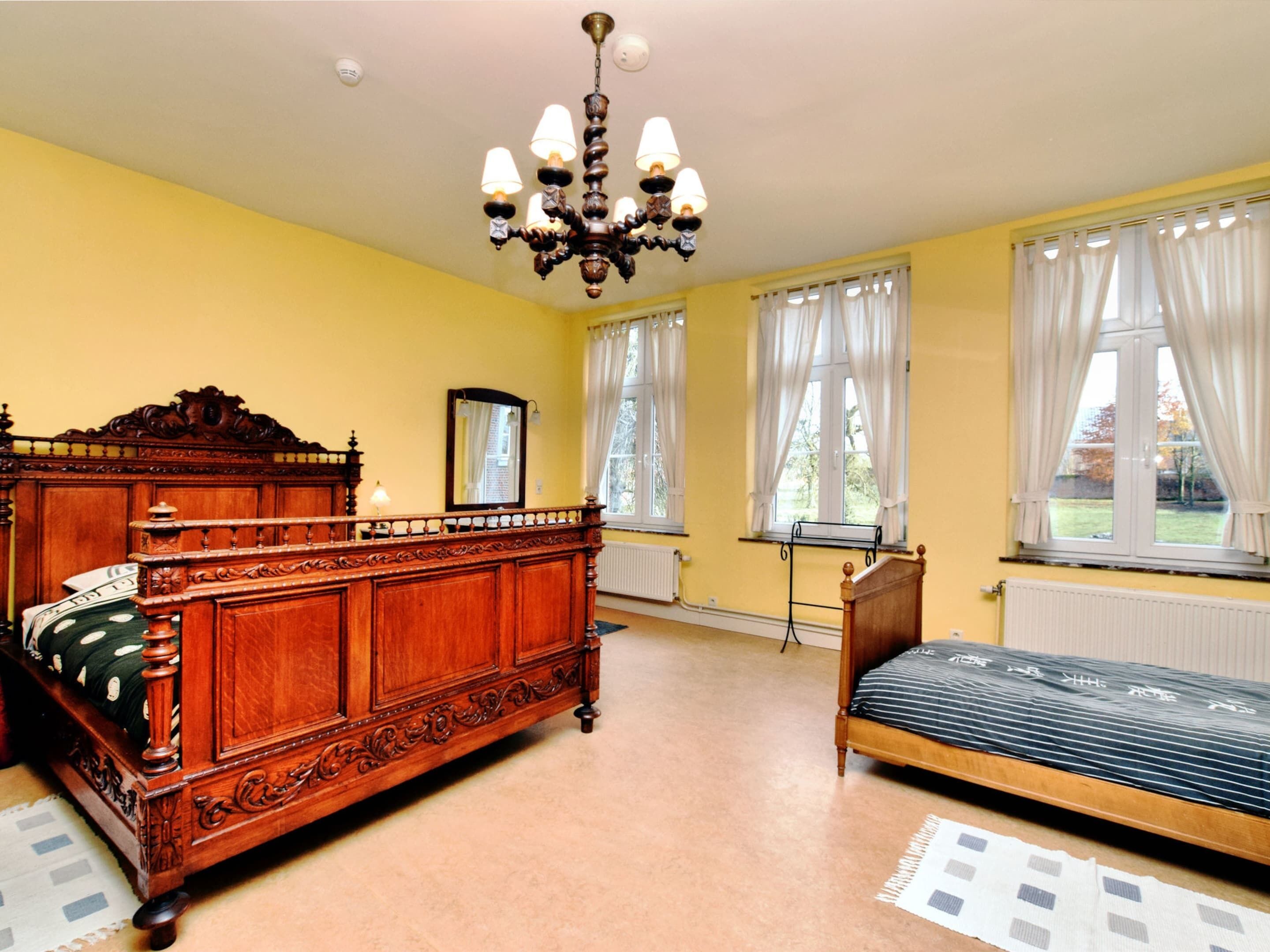 A bedroom with a large wooden bed, a smaller bed, a chandelier, and two windows with curtains.