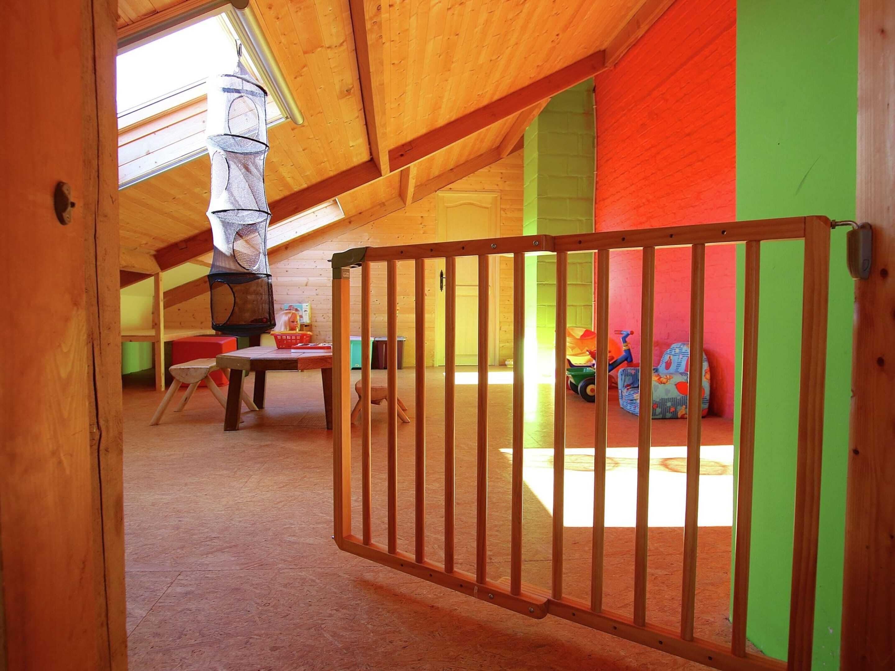 Une pièce aux hauts plafonds, avec un plafond en bois, un parc pour enfants doté d’un sommet en treillis, une table avec des chaises et un espace de jeux coloré rempli de jouets.