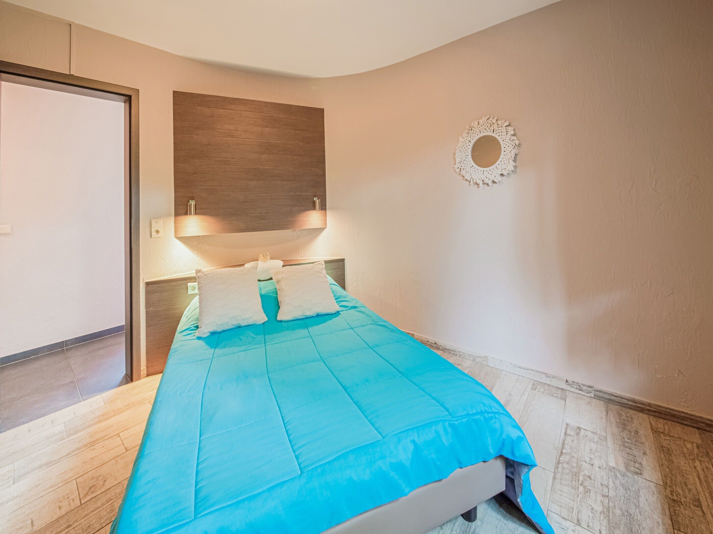 Una cama bien hecha con una colcha azul y dos almohadas, un cabecero de madera, un espejo en la pared y una puerta a la izquierda.