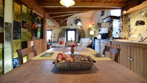 A wooden dining table with a woven basket containing apples and nuts, a vase with flowers, and a view into a kitchen and living area with stone walls and wooden beams. - Holiday Home in Longueville Near Forest (Durbuy)