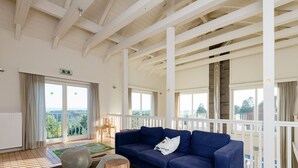 Une pièce spacieuse avec un canapé bleu, des tabourets en bois et une vue sur l’extérieur grâce à de grandes fenêtres.