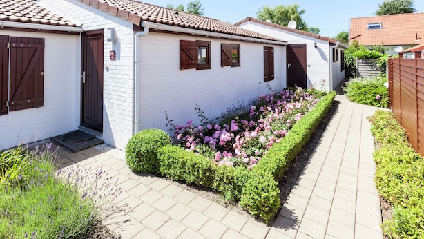 Eine Reihe weißer Gebäude mit braunen Fensterläden, ein gepflasterter Weg und ein Blumenbeet mit rosa und violetten Blumen.