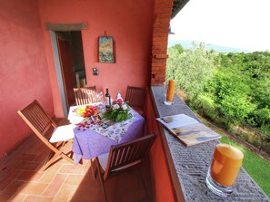 A table set with a purple tablecloth, wine, fruit, and candles on a balcony with a brick wall and a view of greenery.
