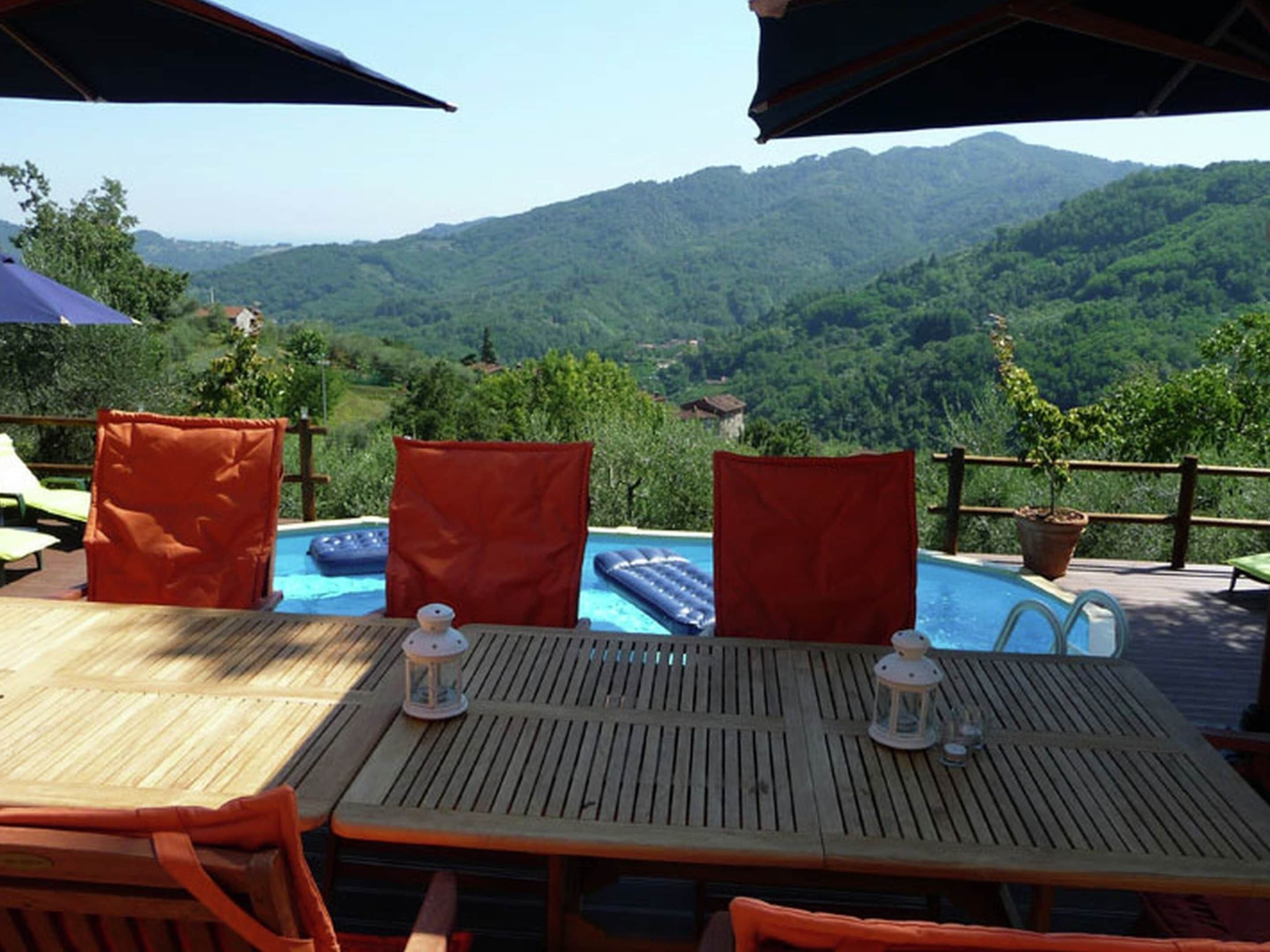 Un área junto a la piscina con mesas y sillas de madera, una piscina flotante y vistas a un paisaje montañoso.