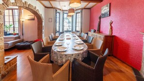 A dining area with a long table set for six, surrounded by wicker chairs, a stone archway, and a red wall with a textured pattern. - Ludogite: Lover's Retreat (Waimes)