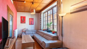 A modern living room with a sectional sofa, a wooden coffee table, a television, and large windows with plants. - Ludogite: Lover's Retreat (Waimes)