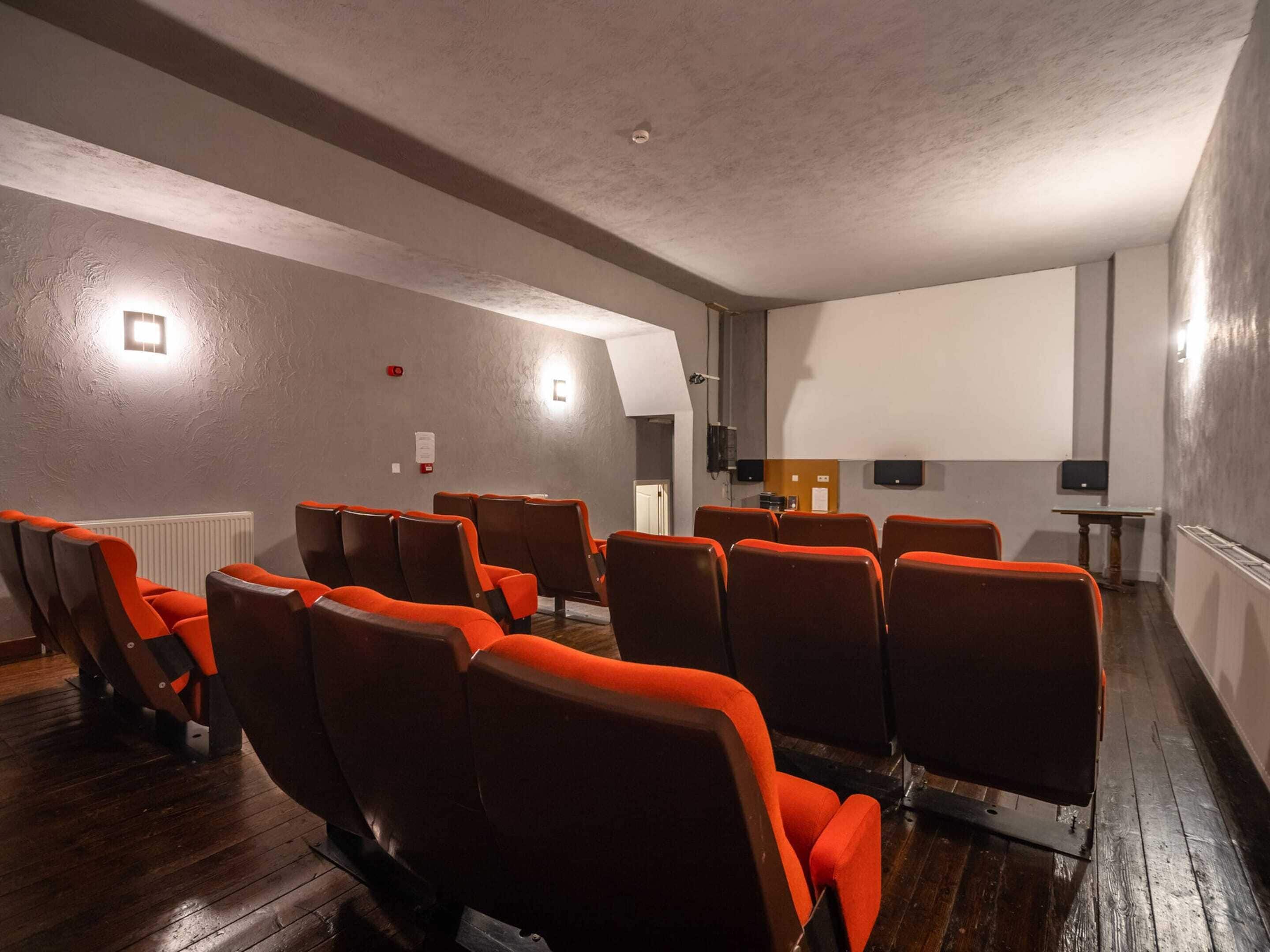 Ein Kinosaal mit Reihen orangener Sitze, einer weißen Leinwand und einem Holzboden.