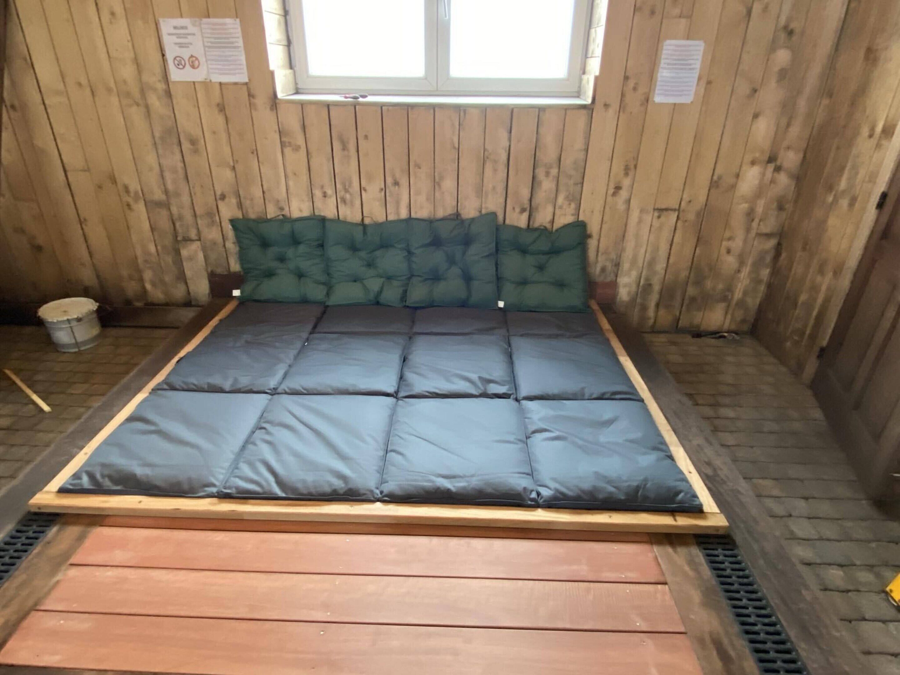 Une pièce aux murs lambrés, avec un lit, une fenêtre et un seau.