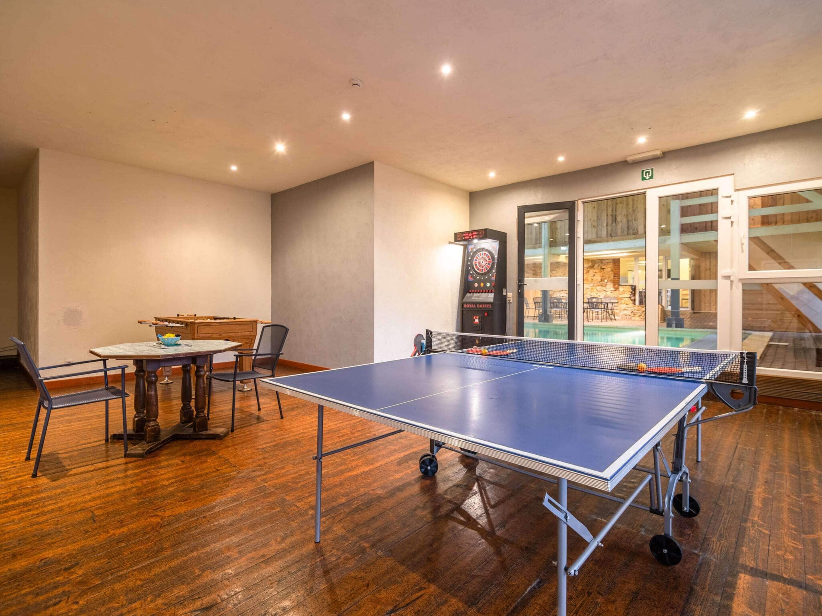 A ping pong table in a room with wooden flooring, a dartboard, and a view of an outdoor swimming pool through glass doors.