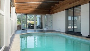 Piscina interior moderna com vigas de madeira no teto e portas de vidro que dão para um terraço com mobiliário de exterior.
