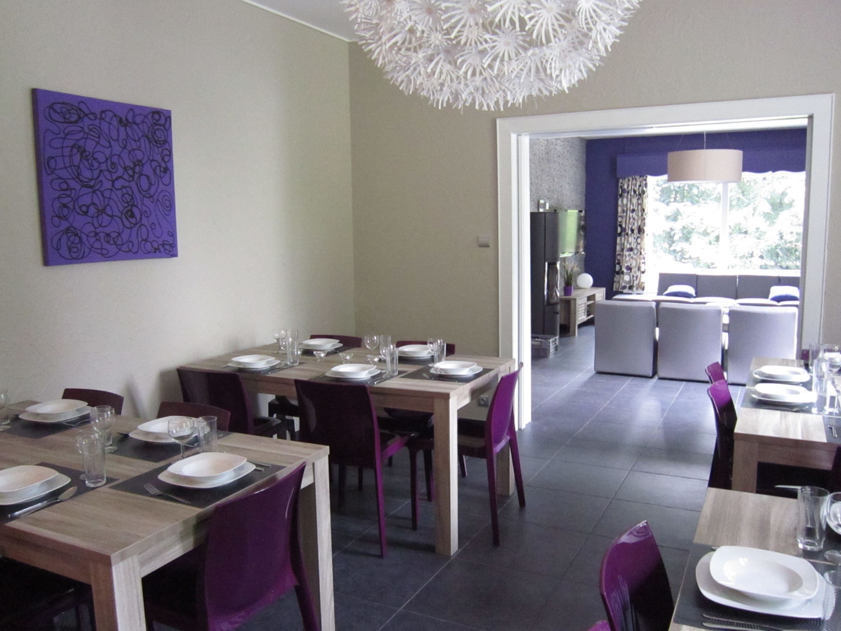 A dining area with a modern design, featuring a wooden table set for four with purple chairs. There is a purple abstract painting on the wall and a white chandelier above the table.