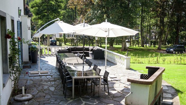 An outdoor patio with stone flooring, a long dining table, chairs, and two white umbrellas. - Luxurious Countryside Holiday Home With Pool (Malmedy)