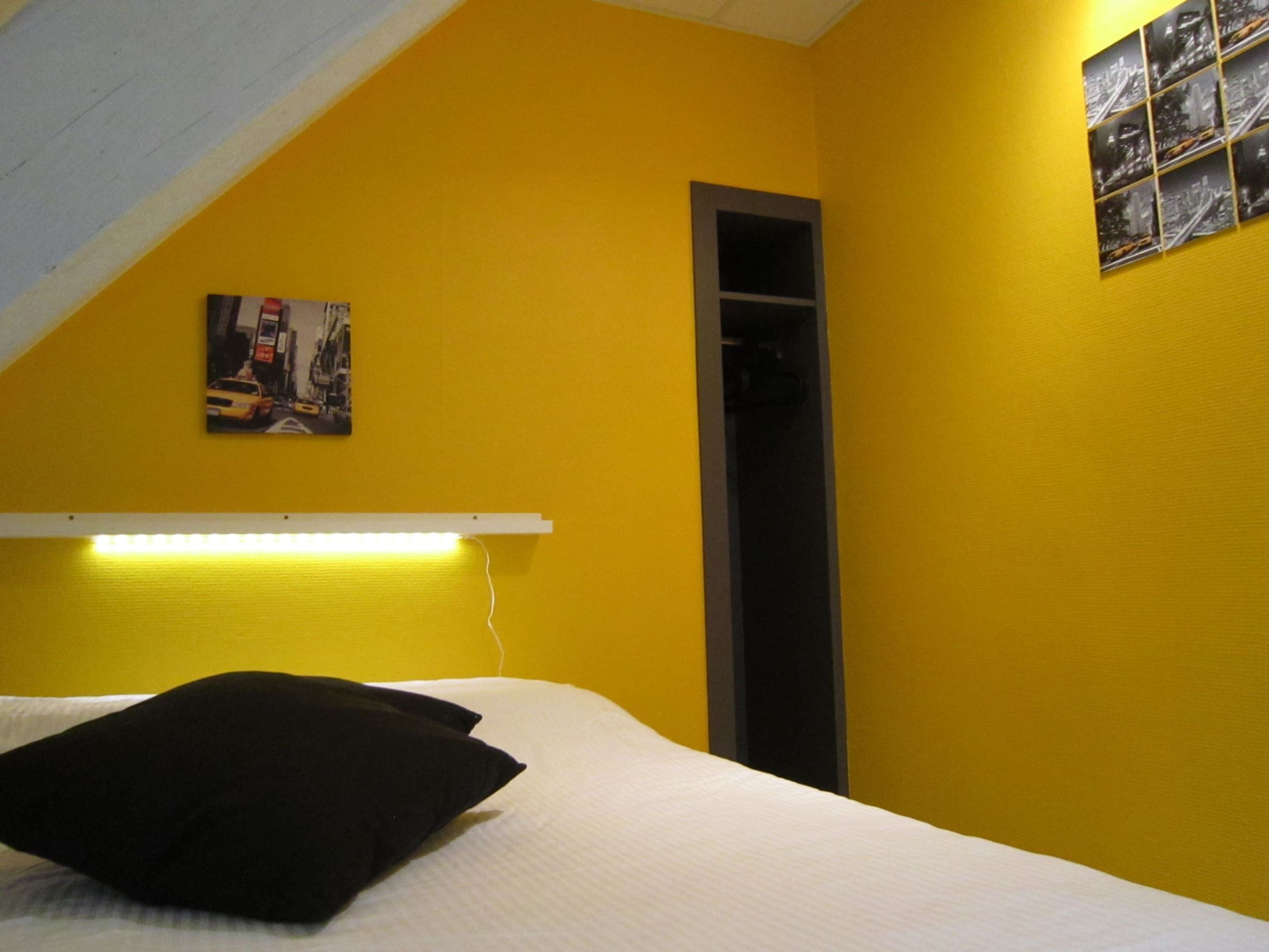A room with a bed, a black pillow, a white headboard, a wall-mounted shelf with a picture, and a vertical black storage unit.