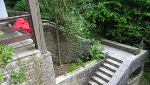 Un escalier en pierre mène à une terrasse en bois agrémentée d’une balustrade, le tout entouré de verdure et d’un mur de briques.