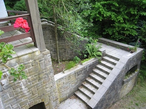 Un escalier en pierre mène à une terrasse en bois agrémentée d’une balustrade, le tout entouré de verdure et d’un mur de briques.
