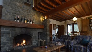 Un salon chaleureux avec une cheminée en pierre, des poutres en bois, des meubles à motifs écossais et une variété de bouteilles disposées sur une étagère.