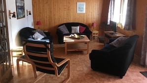 Un salon chaleureux avec des boiseries, un canapé noir, une chaise en bois, une petite table avec un plateau et un téléviseur posé sur un meuble en bois.