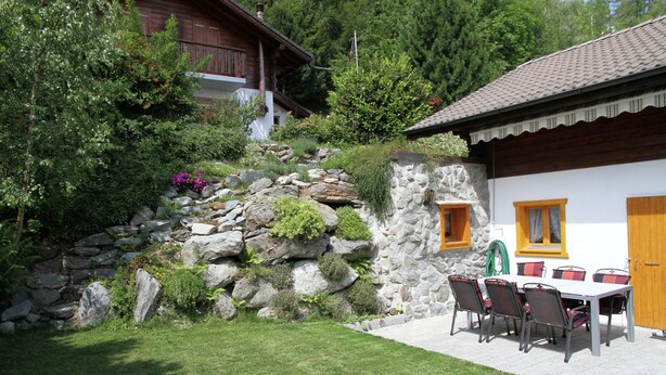 Un mur de pierre bordé d’un jardin, un bâtiment en bois avec un toit en tuiles et une table avec des chaises à l’extérieur.