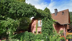 Una casa tradicional con fachada de ladrillo, techo rojo y un jardín con plantas en macetas y un árbol grande.