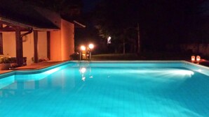 Une piscine nocturne à l’eau claire, entourée d’un bâtiment doté d’un patio couvert et de deux lampes illuminées.