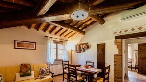 A cozy living room with a wooden ceiling, a dining table with chairs, a sofa with cushions, a radiator, and a window with curtains. - Scenic Farmhouse With Pool (Citta della Pieve)
