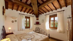 A bedroom with a wooden ceiling, two windows with curtains, a bed with a patterned bedspread, a bedside lamp, and a small shelf with a red item. - Scenic Farmhouse With Pool (Citta della Pieve)