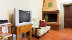 A living room with a television on a wooden stand, a sofa with floral upholstery, and a brick fireplace. - Belvilla by OYO Vintage Holiday Home in Montaione (Montaione)