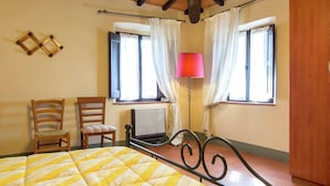 A bedroom with a bed, two chairs, a radiator, and two windows with curtains. - Belvilla by OYO Vintage Holiday Home in Montaione (Montaione)