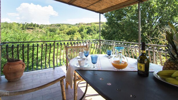 A covered outdoor dining area with a table set for two, a bottle of wine, and a plate of bananas. - Belvilla by OYO Vintage Holiday Home in Montaione (Montaione)