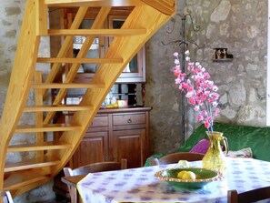 Uma escada de madeira leva a uma cozinha com parede de pedra, uma mesa de jantar com toalha florida e um vaso com flores cor-de-rosa.