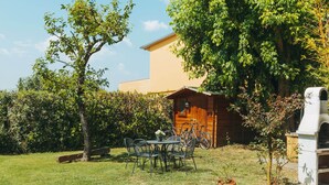 A wooden cabin with a table and chairs in a grassy area, surrounded by trees and a building in the background. - Belvilla by OYO Gli Archi (Cortona)