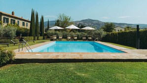 A rectangular swimming pool with a clear blue water, surrounded by a well-maintained lawn and a stone border. There are lounge chairs and umbrellas for shade, and a building with a tiled roof in the background. - Belvilla by OYO Gli Archi (Cortona)