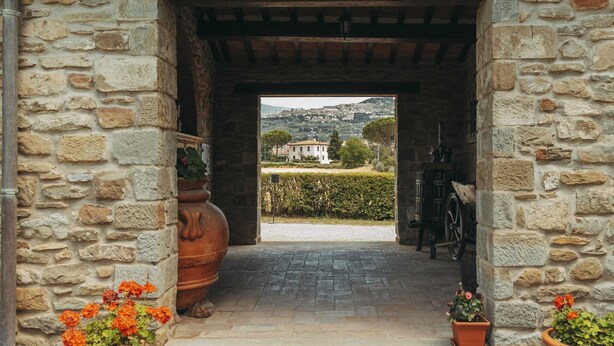 A stone archway leading to a paved courtyard with potted plants and a view of a distant building. - Belvilla by OYO Gli Archi (Cortona)