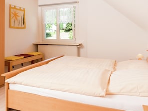 A bedroom with a bed, a window, a radiator, and a small table with a cushion.