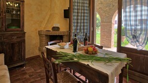 Un coin repas avec une table en bois dressée pour deux personnes, une cheminée en briques et une vue sur le jardin par une fenêtre agrémentée de rideaux.