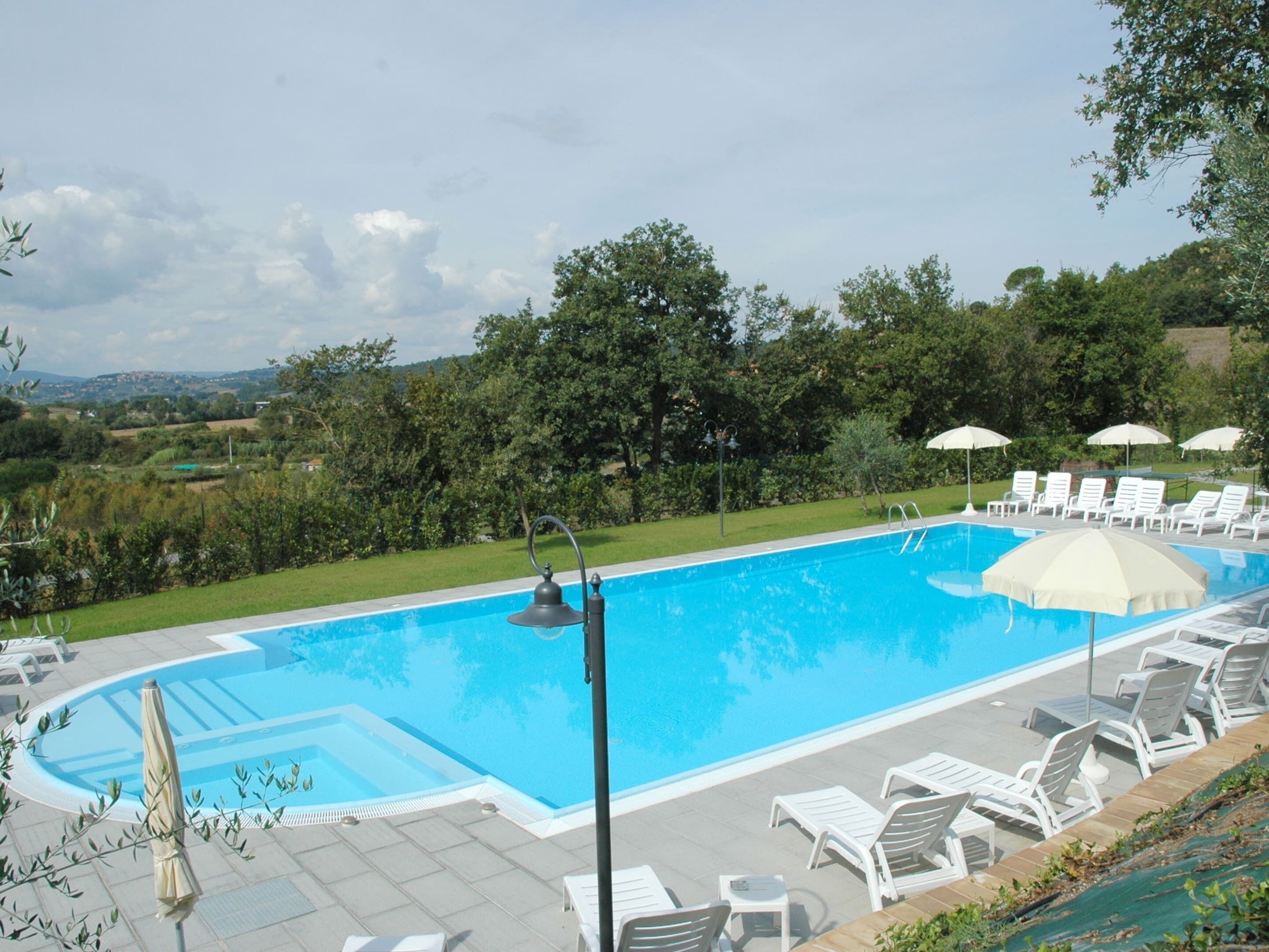A swimming pool with lounge chairs and umbrellas, surrounded by greenery and a view of the countryside.