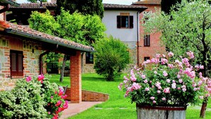 A garden with a stone building, a wooden barrel planter with pink flowers, and a brick house in the background. - Villa in Citta Della Pieve Near Lake (Citta della Pieve)