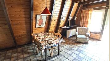 Um quarto de sótão aconchegante com teto inclinado de madeira, uma mesa de jantar com toalha de mesa estampada, uma cadeira, uma televisão e uma janela com cortinas.