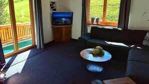 Een woonkamer met een bank, een bijzettafeltje met bloemen, een televisie op een houten dressoir, en grote ramen met gordijnen.