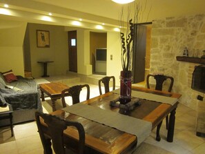 Ein Essbereich mit Holztisch, Stühlen und einer Vase mit Zweigen. Im Hintergrund befindet sich eine Couch und ein Kamin.