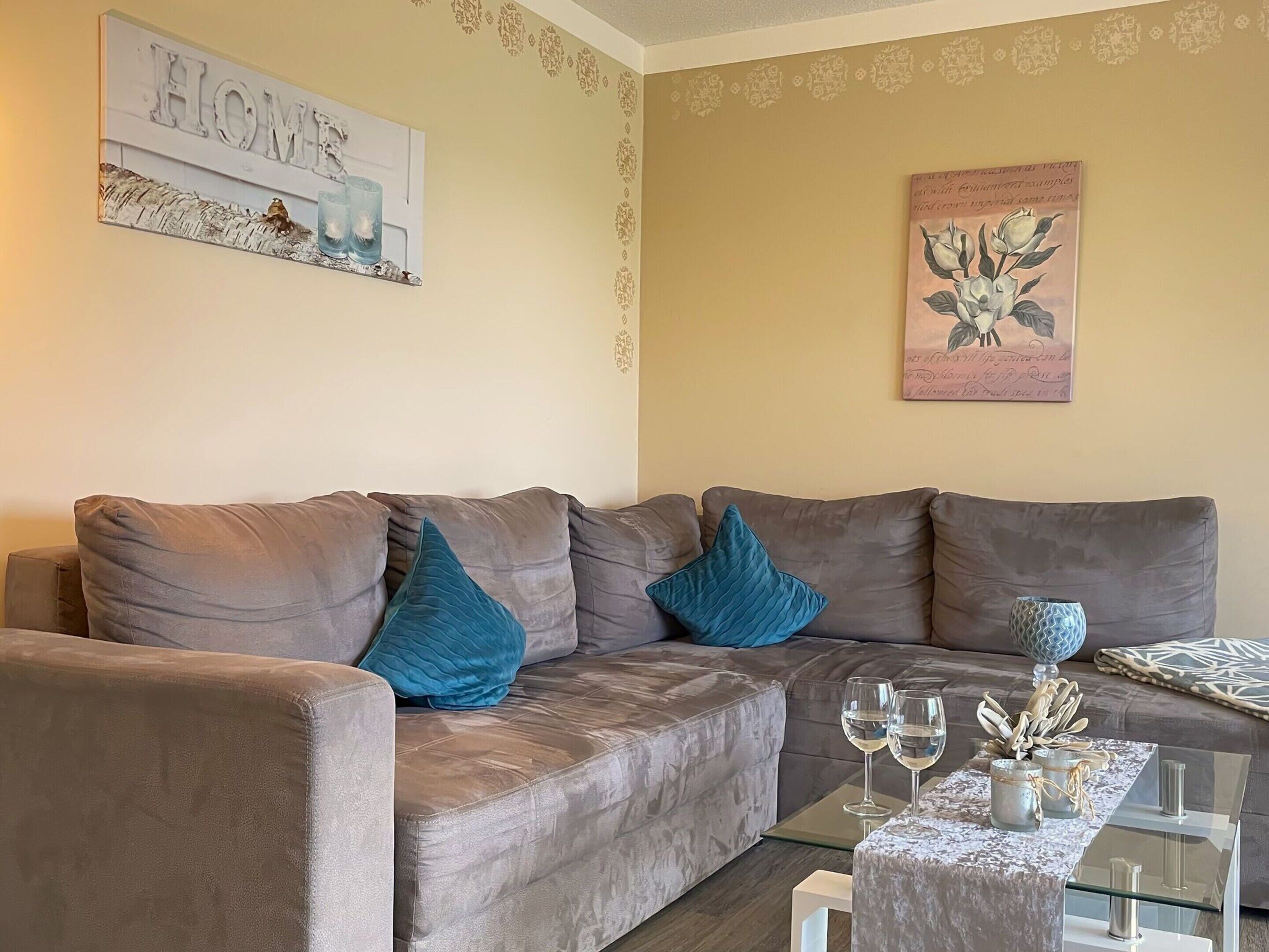 Una sala de estar con un sofá gris, dos cojines azules, una mesa de centro de vidrio con copas de vino y objetos decorativos, y detalles de decoración en la pared, incluyendo un letrero que dice 'HOME' y una pintura floral.