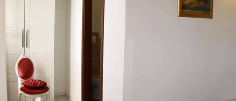 A room with a white wardrobe, a red cushioned chair, a small table, and a painting on the wall.