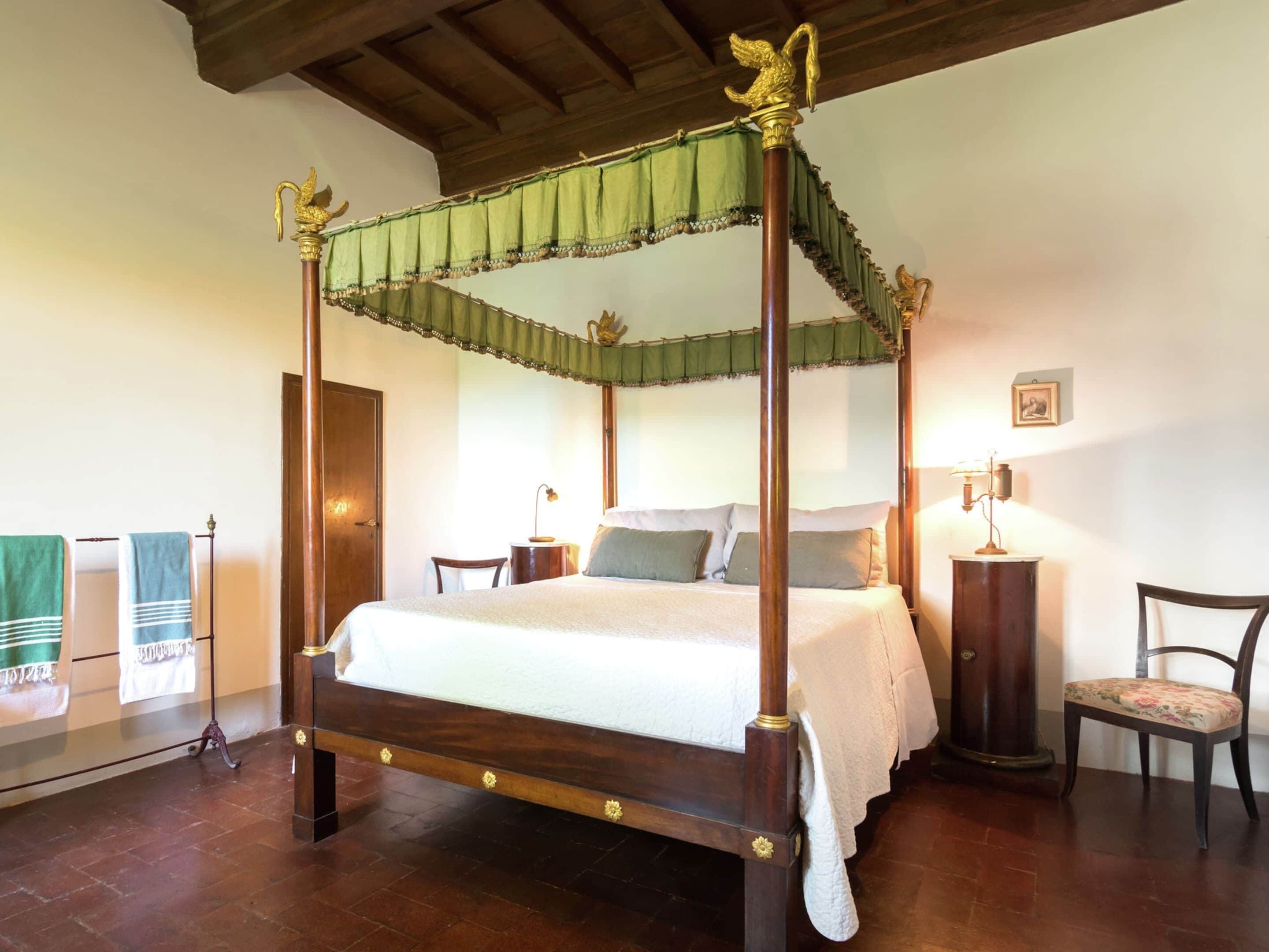 Un letto a baldacchino con tenda verde, pavimento in legno, una sedia, un comodino e un quadro appeso al muro.
